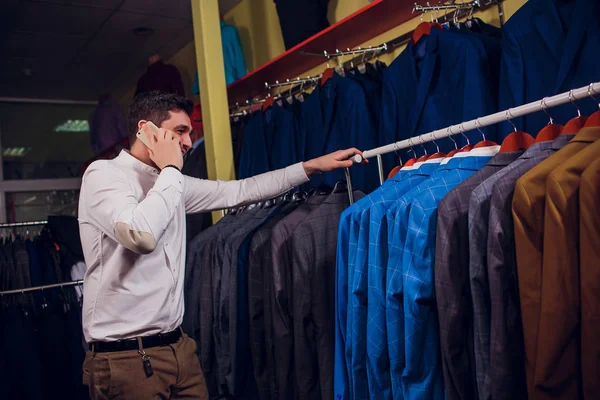sale, shopping, fashion, communication and people concept - happy young man or businessman calling on smartphone and choosing clothes clothing store
