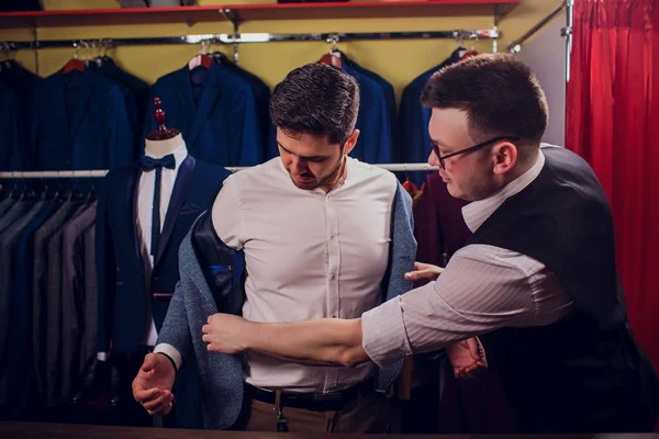 Empresario en chaleco clásico contra los trajes de fila en la tienda. El hombre ayuda a probar otro traje en la tienda de ropa — Foto de Stock
