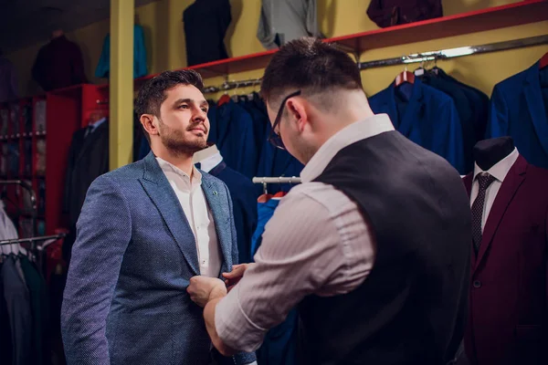 Empresario en chaleco clásico contra los trajes de fila en la tienda. El hombre ayuda a probar otro traje en la tienda de ropa —  Fotos de Stock