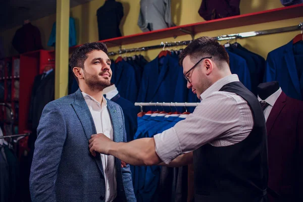 Uomo d'affari in giubbotto classico contro abiti da fila in negozio. L'uomo aiuta un'altra prova sul vestito nel negozio di abbigliamento — Foto Stock