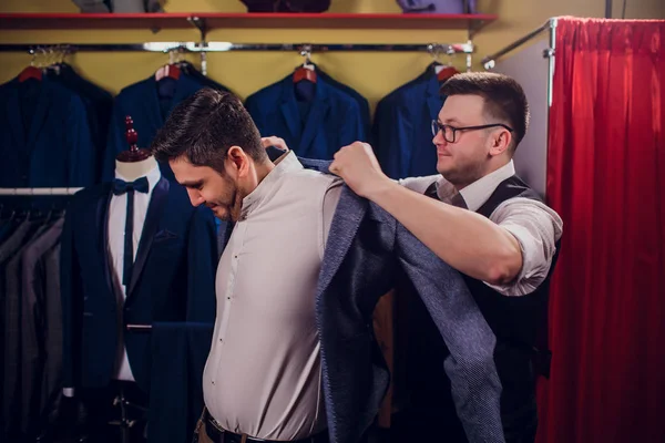 Empresario en chaleco clásico contra los trajes de fila en la tienda. El hombre ayuda a probar otro traje en la tienda de ropa — Foto de Stock