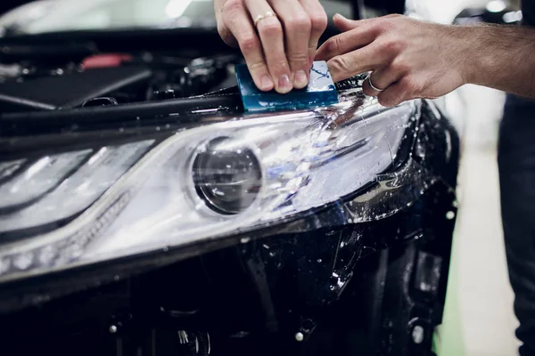 Worker hands installs car paint protection film wrap auto headlight