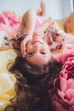 Studio iç çiçeklerle süslenmiş oturan şık elbiseli çekici genç kadın. Güzel taze gülümseyen kız çiçekleri ile poz