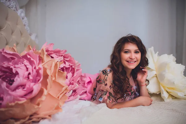 Jovem atraente em vestido elegante sentado em estúdio interior decorado com flores. Menina sorridente fresco bonito posando com flores — Fotografia de Stock