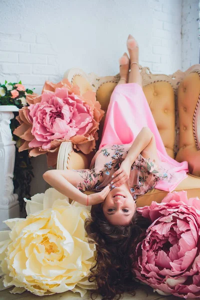 Aantrekkelijke jonge vrouw in elegante jurk zitten in studio interieur versierd met bloemen. Mooi vers lachende meisje poseren met bloemen — Stockfoto