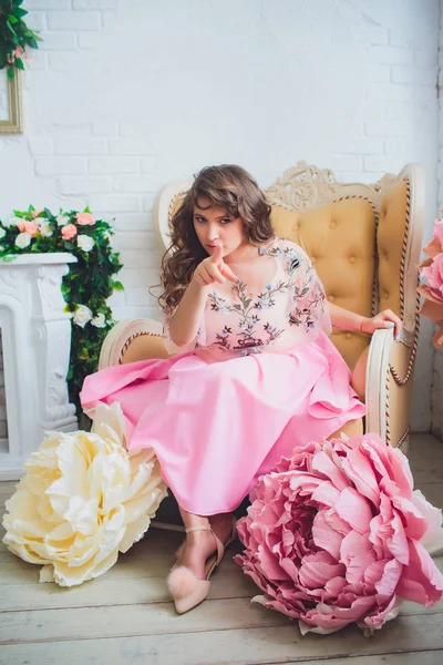 Jovem atraente em vestido elegante sentado em estúdio interior decorado com flores. Menina sorridente fresco bonito posando com flores — Fotografia de Stock