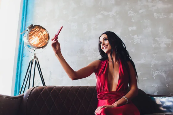 Foto de estudio de moda de mujer hermosa con cabello oscuro y maquillaje de noche, lleva vestido rojo de lujo. luces turquesa neón proyector de película hace una selfie — Foto de Stock