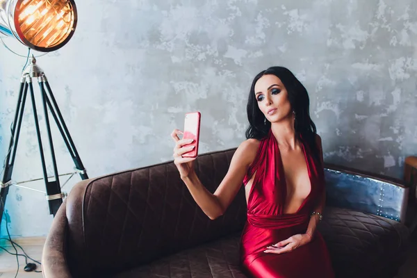 Foto estúdio de moda de mulher linda com cabelo escuro e maquiagem à noite, usa vestido vermelho luxuoso. luzes neon turquesa holofotes filme faz uma selfie — Fotografia de Stock