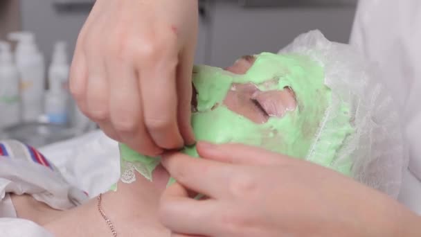 Cosmetologist με τη βοήθεια ενός πινέλου ειδικό μακιγιάζ κάνει μια μάσκα ΙΑΜΑΤΙΚΟΣ από πράσινα φύκια κατά της γήρανσης του μια νεαρή γυναίκα. — Αρχείο Βίντεο