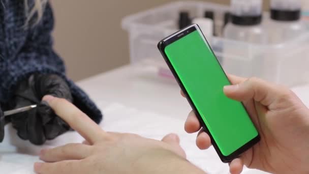 Smartphone de pantalla verde. hombre desplazándose por las redes sociales utilizando el teléfono moderno de pantalla táctil agradable. Manicura en salón de belleza. Ella espera que el esmalte de uñas se seque en su brazo . — Vídeos de Stock