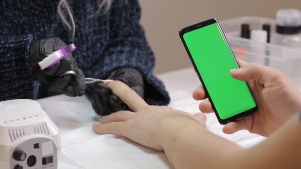 Smartphone de pantalla verde. hombre desplazándose por las redes sociales utilizando el teléfono moderno de pantalla táctil agradable. Manicura en salón de belleza. Ella espera que el esmalte de uñas se seque en su brazo . — Vídeos de Stock