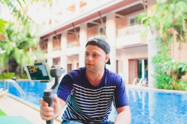 Genç erkek video Blogger onun kanalı için video içeriği oluşturur. Havuz kenarında otururken kullanıcılar için video çekimleri. Adam vlogger sabitleyici ile kamera kendini rahatlatır.