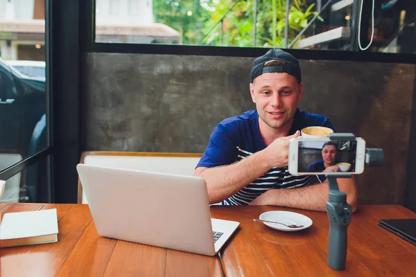 Bearded attractive male video blogger in stylish glasses shoots video streaming for users while sitting in cafe and drinking coffee. Male vlogger takes pictures of himself on camera with stabilizer.