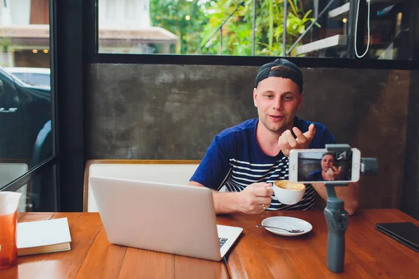 Bearded attractive male video blogger in stylish glasses shoots video streaming for users while sitting in cafe and drinking coffee. Male vlogger takes pictures of himself on camera with stabilizer.