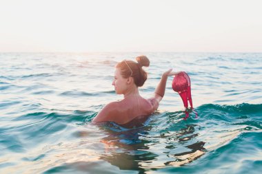 Uzun saçlı Genç kadın, Sarışın, topless, suda oturan ve elinde tutan güneşte bir bikini üst.