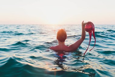Uzun saçlı Genç kadın, Sarışın, topless, suda oturan ve elinde tutan güneşte bir bikini üst.