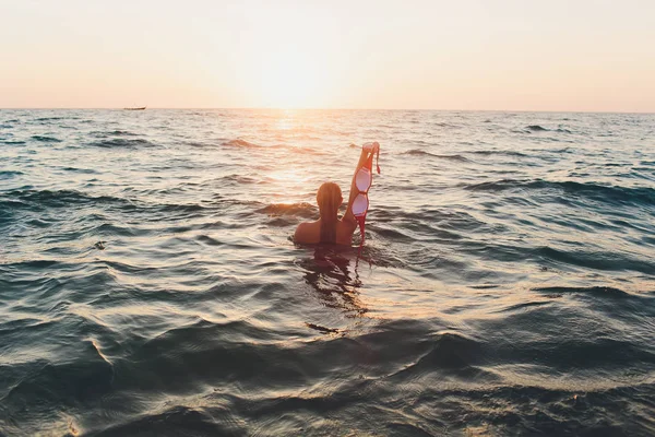 Jeune femme aux cheveux longs, blonde, topless, assise dans l'eau et tenant dans sa main un haut de bikini au soleil . — Photo