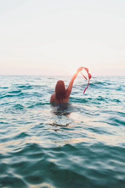 Jonge vrouw met lang haar, blond, topless, zittend in het water en holding in zijn hand een bikini top in de zon. — Stockfoto