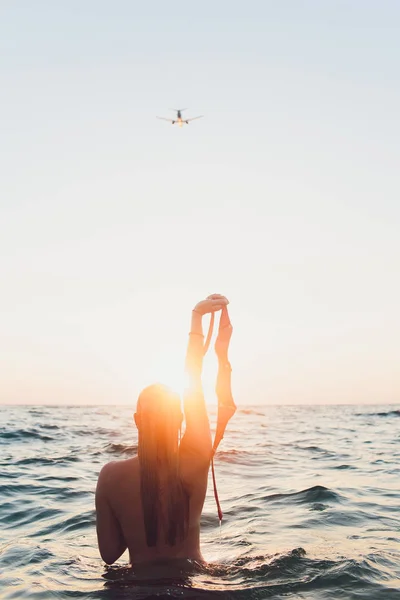 Jonge vrouw met lang haar, blond, topless, zittend in het water en holding in zijn hand een bikini top in de zon. nam haar beha, vliegtuig, zee — Stockfoto