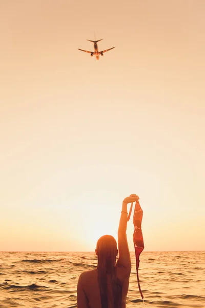 Young woman with long hair, blonde, topless, sitting in the water and holding in his hand a bikini top in the sunshine. took off her bra, plane, sea