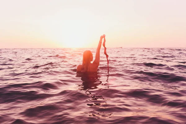 Jonge vrouw met lang haar, blond, topless, zittend in het water en holding in zijn hand een bikini top in de zon. — Stockfoto