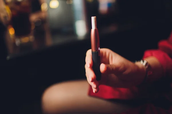 Sigarette elettroniche, sigarette tecnologiche. Sistema tabacco IQOS. Mano femminile su sfondo sfocato del ristorante. caso rosso . — Foto Stock