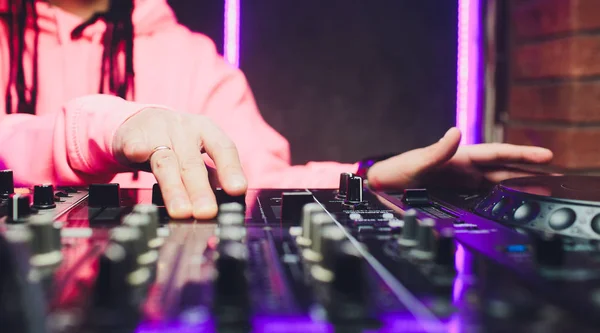 Close up view of the hands of male disc jockey mixing music on his deck with his hands poised over the vinyl record on the turntable and the control switches at night.
