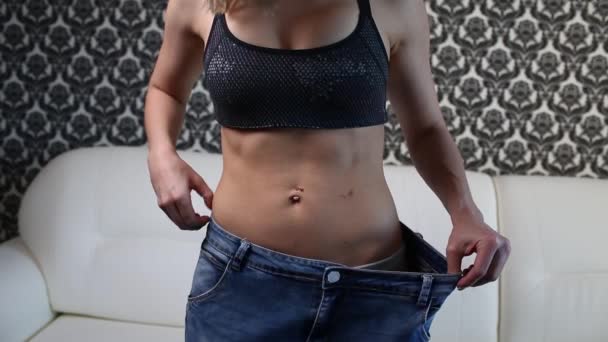 Young woman shows her weight loss and wearing her old jeans at home in light bedroom. A slim girl in too big jeans showing how she was losing weight when she started eating healthy food. — Stock Video