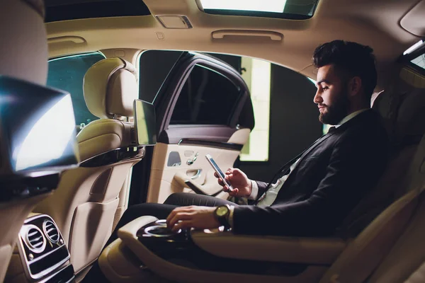 Día de trabajo ocupado. Hombre joven guapo en traje completo mirando su teléfono inteligente mientras está sentado en el coche . — Foto de Stock