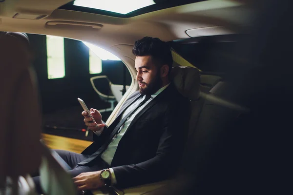 Mozgalmas munkanap. Csinos fiatalember teljes öltönyt nézi okos telefon ülve az autóban. — Stock Fotó