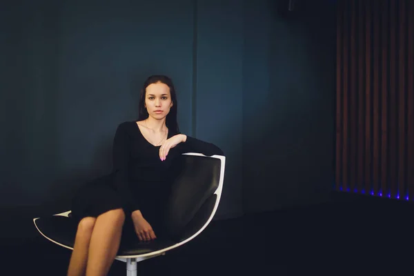 現代的な肘掛け椅子に座っている黒いカクテルドレスとハイヒールのエレガントな美しい女性. — ストック写真
