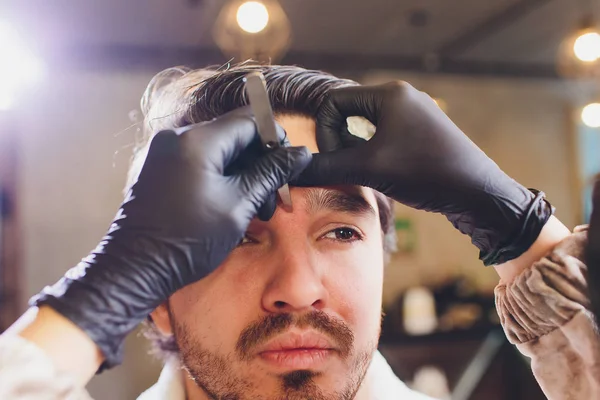 Closeup of process of threading procedure in barber shop. Professional barber correcting shape of brows with threads to smiling young man sitting in chair. Concept of eyebrows care.