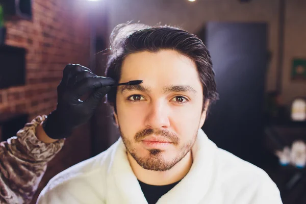 Closeup of process of threading procedure in barber shop. Professional barber correcting shape of brows with threads to smiling young man sitting in chair. Concept of eyebrows care.