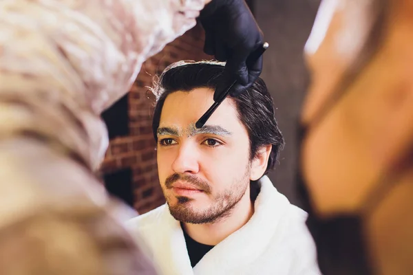 Closeup of process of threading procedure in barber shop. Professional barber correcting shape of brows with threads to smiling young man sitting in chair. Concept of eyebrows care.