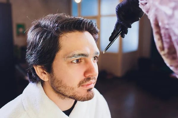 Closeup of process of threading procedure in barber shop. Professional barber correcting shape of brows with threads to smiling young man sitting in chair. Concept of eyebrows care.