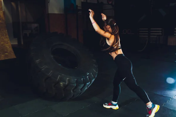 В спортзале фитнесс-женщина перевернула колесо. Подходящая спортсменка, тренирующаяся с огромной шиной. Вид сзади. Женщина занимается силовыми упражнениями . — стоковое фото