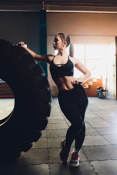 Mulher fitness lançando pneu de roda no ginásio. Apto atleta feminino trabalhando com um pneu enorme. Vista traseira. Desportista fazendo um treinamento de exercícios de força . — Fotografia de Stock