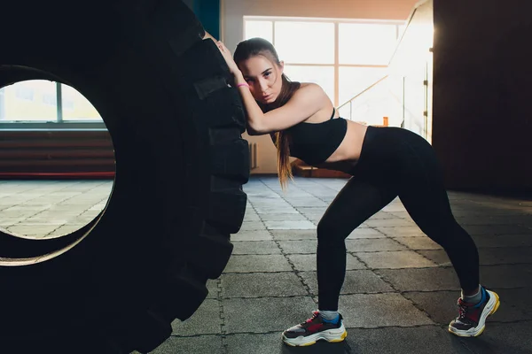 Mujer fitness volteando neumático de rueda en el gimnasio. Ajuste atleta femenina haciendo ejercicio con un neumático enorme. Vista trasera. Deportiva haciendo un entrenamiento de fuerza . — Foto de Stock