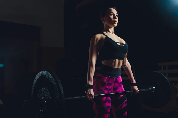 a beautiful sports girl trains a bicep with a rod in her hands in the gym.