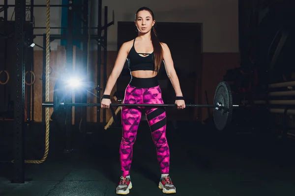 a beautiful sports girl trains a bicep with a rod in her hands in the gym.