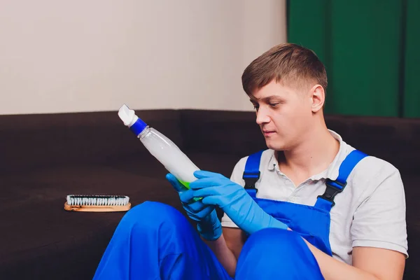 Άνθρωπος να διαβάζει οδηγίες για το μπουκάλι του απορρυπαντικού στο σπίτι, κοντά στον καναπέ του καναπέ. με μπλε προστατευτικά γάντια — Φωτογραφία Αρχείου