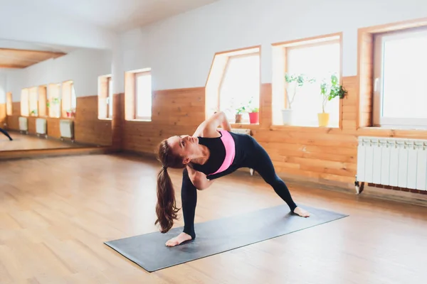 Vista trasera de la joven yogui femenina en sujetador deportivo y polainas haciendo pose de ángulo lateral extendido encuadernado, baddha utthita parsvakonasana, mientras practica yoga en interiores . — Foto de Stock