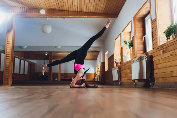 Jeune femme attrayante yogi pratiquant le concept de yoga, debout dans l'exercice de salamba sirsasana, pose de la tête, s'entraîner, porter des vêtements de sport, débardeur noir et pantalon, pleine longueur, fond loft . — Photo