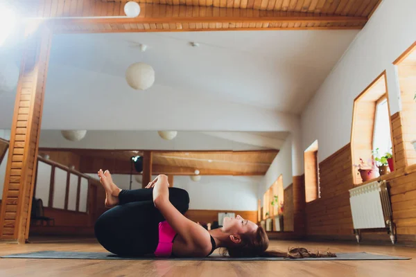 Desportista focada no alongamento antes da aula de Pilates. Menina fazendo aquecimento para as pernas no tapete. Mulher tem cabelo puxado para trás, olhar concentrado. Atleta vestida em top escuro, ioga legging trens em fitness . — Fotografia de Stock