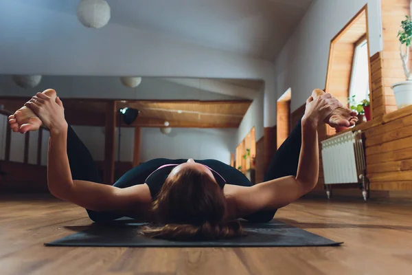 Mujer atractiva joven practicando yoga, estiramiento en ejercicio Ananda Balasana, pose de bebé feliz, entrenamiento, usar ropa deportiva negra, estilo urbano fresco, larga duración, fondo de estudio gris, vista lateral — Foto de Stock