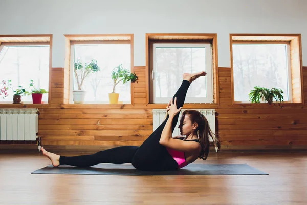Young yogi sporty woman working out, warming up using yogic belt, lying in yoga Supta Padangushthasana, One Leg Lift exercise, reclined variation of Extended Hand to Big Toe pose, studio, side view.