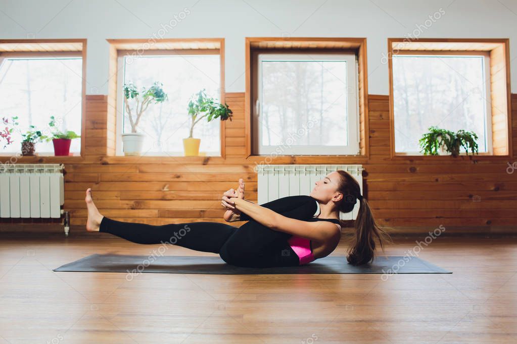 Young yogi sporty woman working out, warming up using yogic belt, lying in yoga Supta Padangushthasana, One Leg Lift exercise, reclined variation of Extended Hand to Big Toe pose, studio, side view.