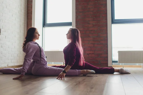 Profesyonel iki kadın sporcu birlikte yoga pozisyonunda oturuyor. Kollarına dokunuyorlar ve birbirlerine dinginlikle bakıyorlar. Büyük pencereden güneş ışığı. — Stok fotoğraf