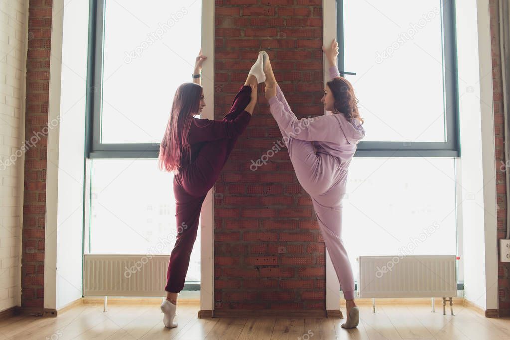 Professional two female athletes are sitting in yoga position together. They are touching their arms and looking at each other with serenity. Sunshine from big window.
