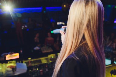 Kız öğrenci sol elinde siyah kablosuz mikrofon tutuyor ve bulanık arka plan ile kamu konuşma yapmak.